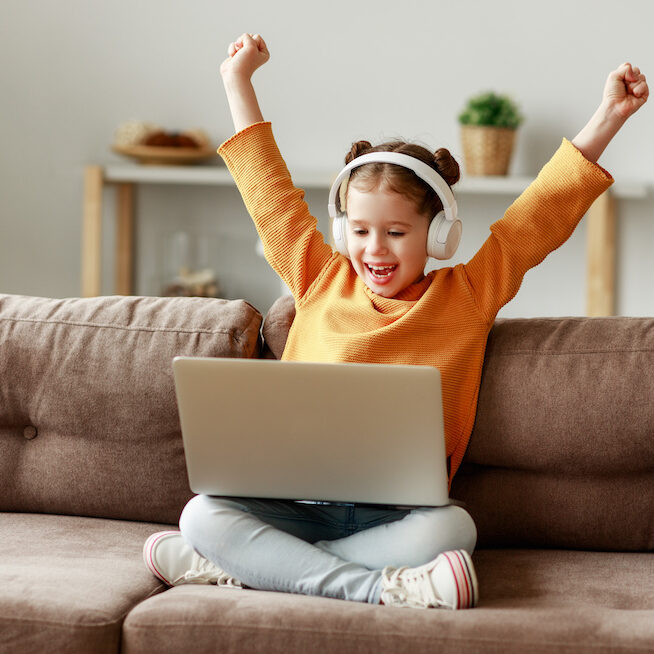 Happy girl in headphones raising arms and smiling while sitting crossed legged on sofa and celebrating victory in video game on laptop